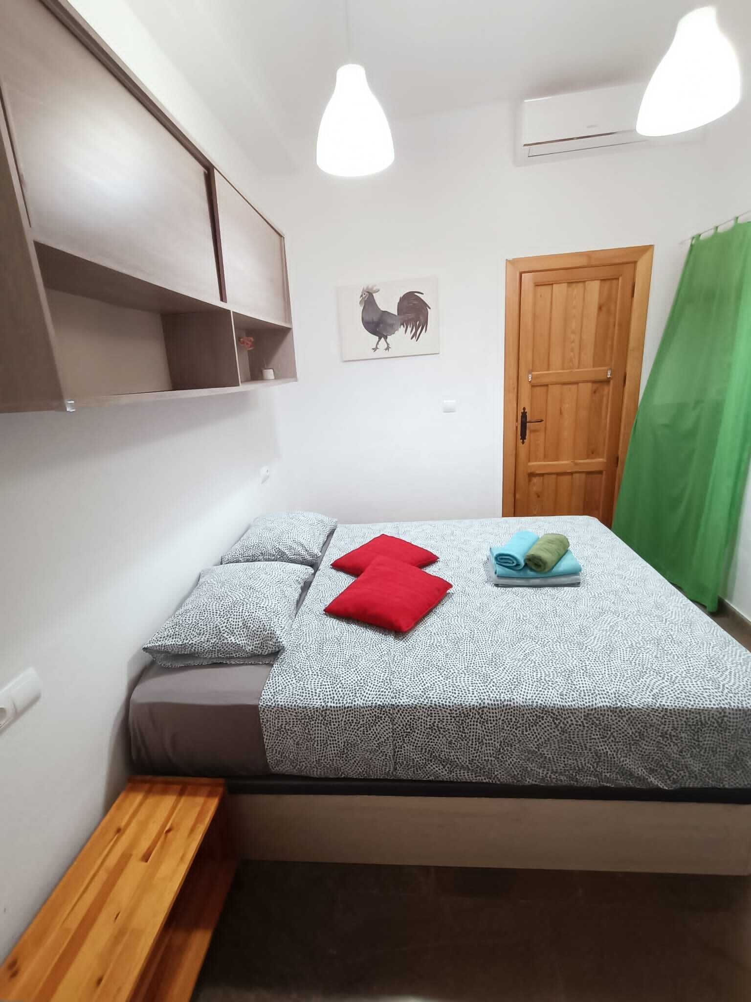 A bedroom with a bed, pillows, a bench, a wooden door, and a wall-mounted shelf.