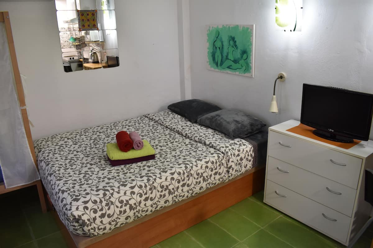 A bedroom with a bed, a TV on a stand, a small desk with drawers, a mirror, and a painting on the wall.