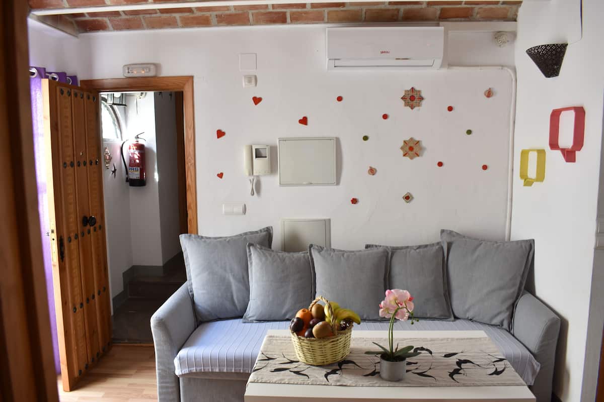 A living room with a sofa, a table with a basket of fruit, a potted plant, and a fire extinguisher on the wall.