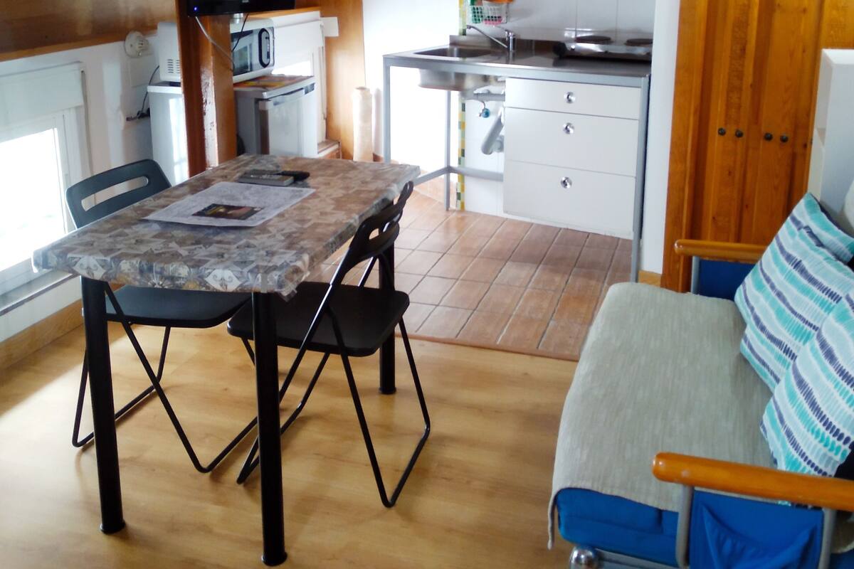 A compact kitchen with a microwave, sink, and stove. There is a dining table with chairs and a sofa with a striped cushion.
