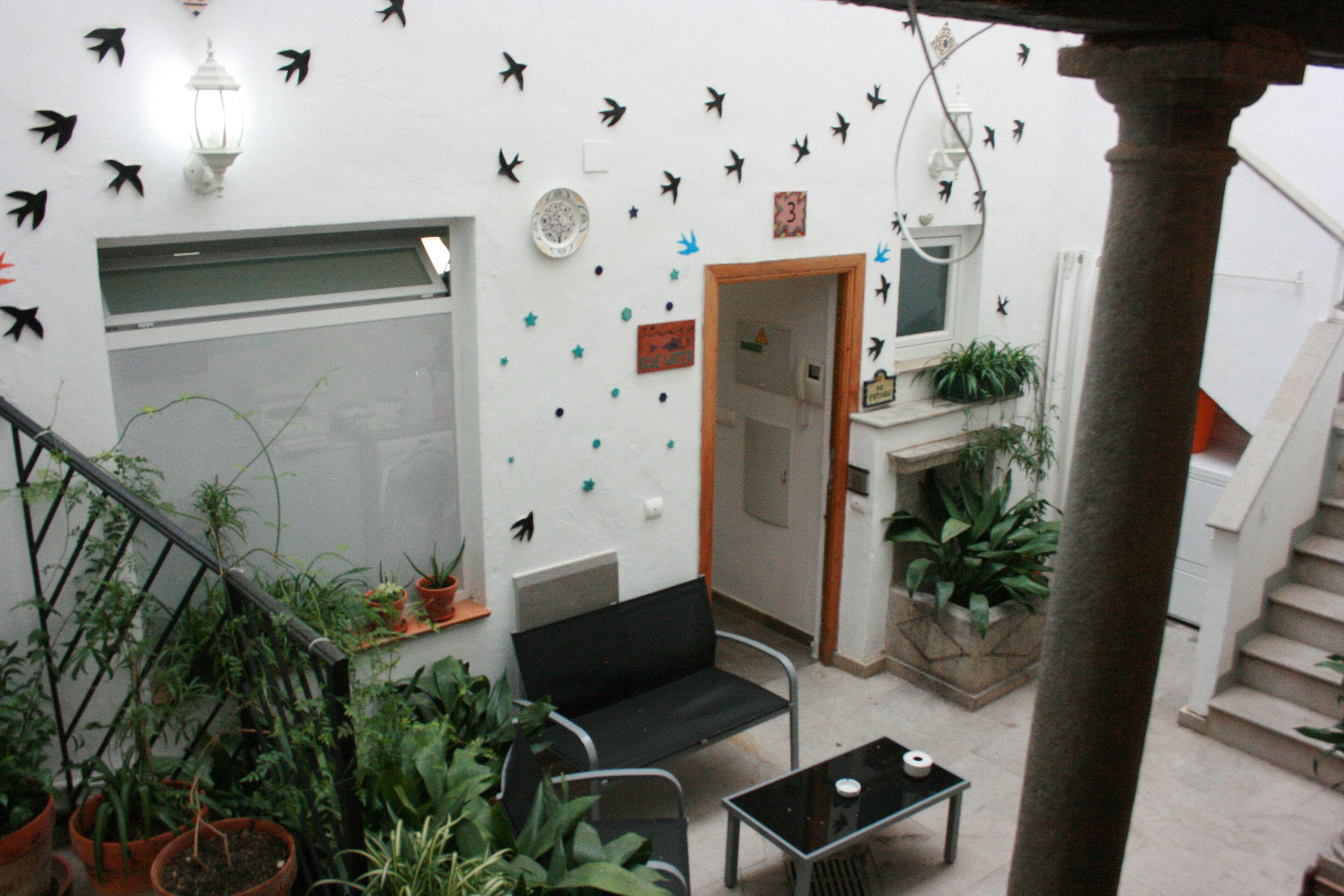 Una habitación con un sofá negro, una mesita y varias plantas en macetas. Se observa una escalera con barandilla y una puerta que da a otra habitación.