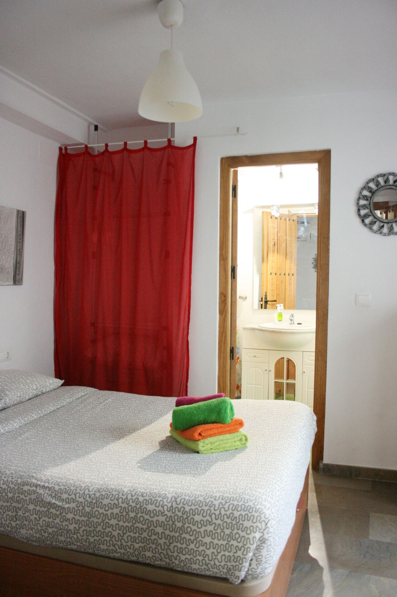 Un dormitorio con una cama, cortinas rojas, una puerta de madera que da a un baño y una lámpara colgante blanca.