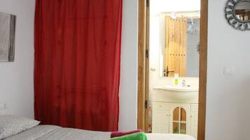 Un dormitorio con una cama, cortinas rojas, una puerta de madera que da a un baño y una lámpara colgante blanca.
