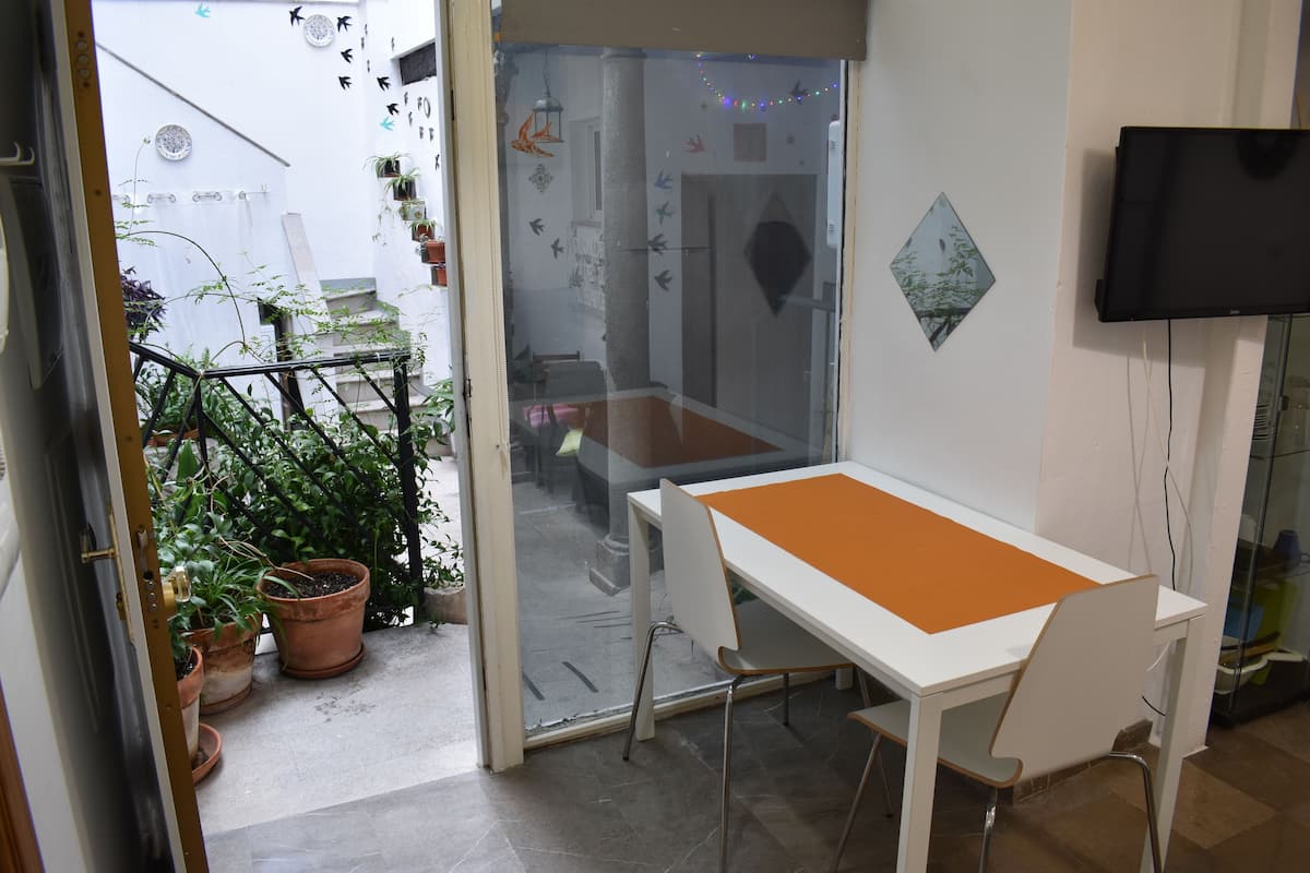 A room with a white table, orange table runner, and a TV mounted on the wall. There are potted plants outside the door.