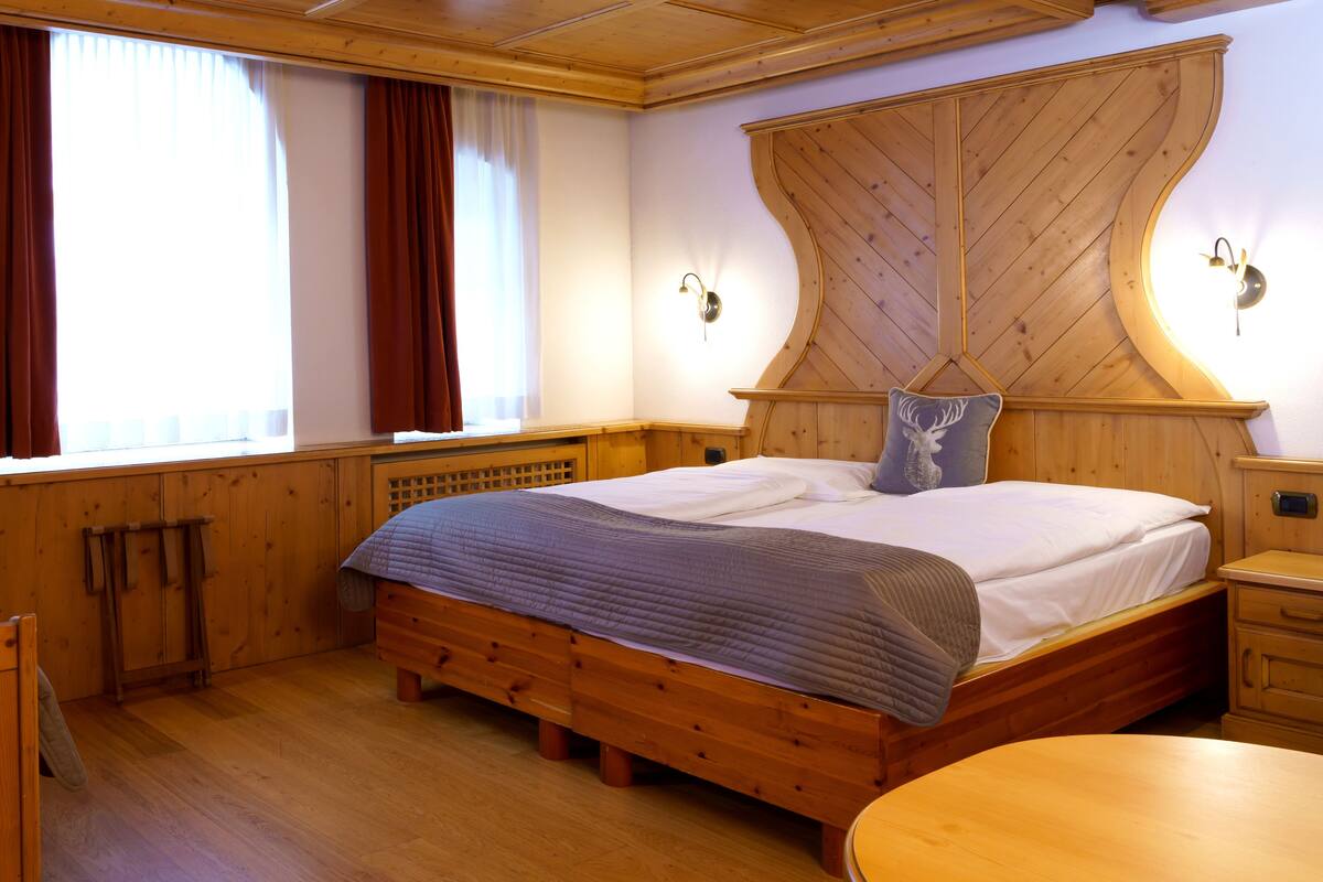 A wooden-framed bedroom with a large bed, a wooden nightstand, a round table, and a window with curtains.