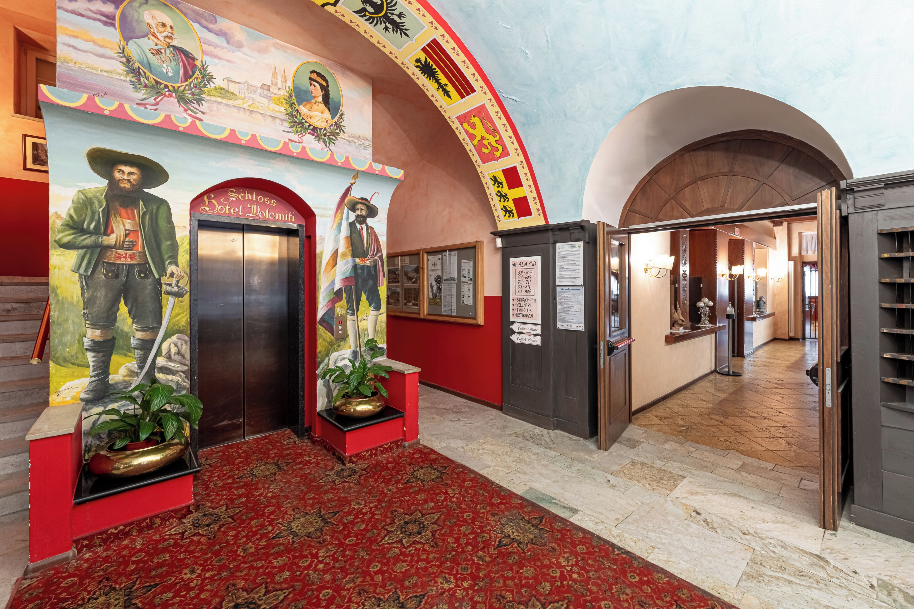 Un hall d’hôtel avec un tapis rouge, deux ascenseurs et une fresque représentant des personnages historiques.