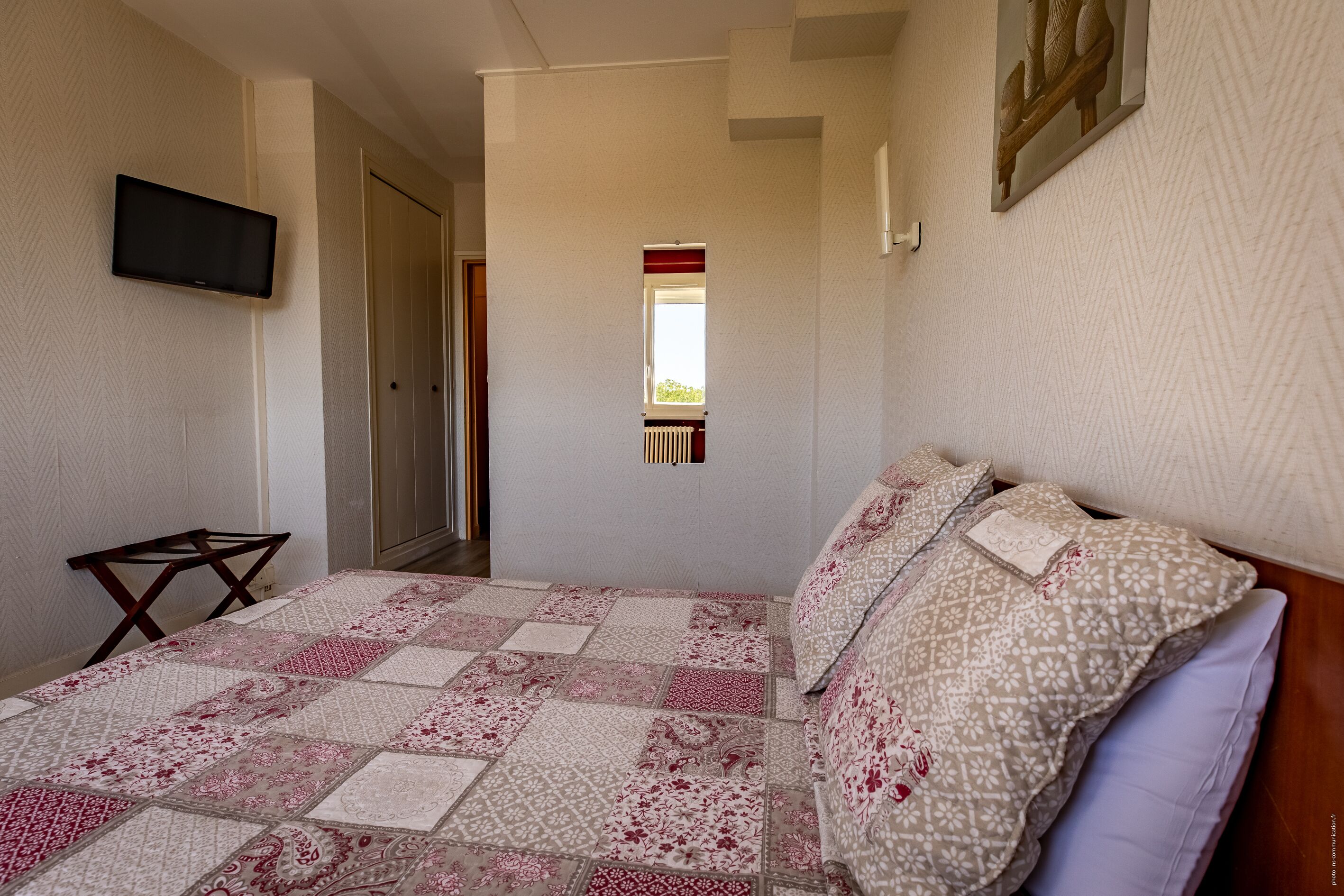 Un dormitorio con una colcha acolchada, un televisor montado en la pared y una ventana con cortina roja.