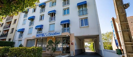 Um hotel com vários andares, varandas, entrada central e área de estacionamento.