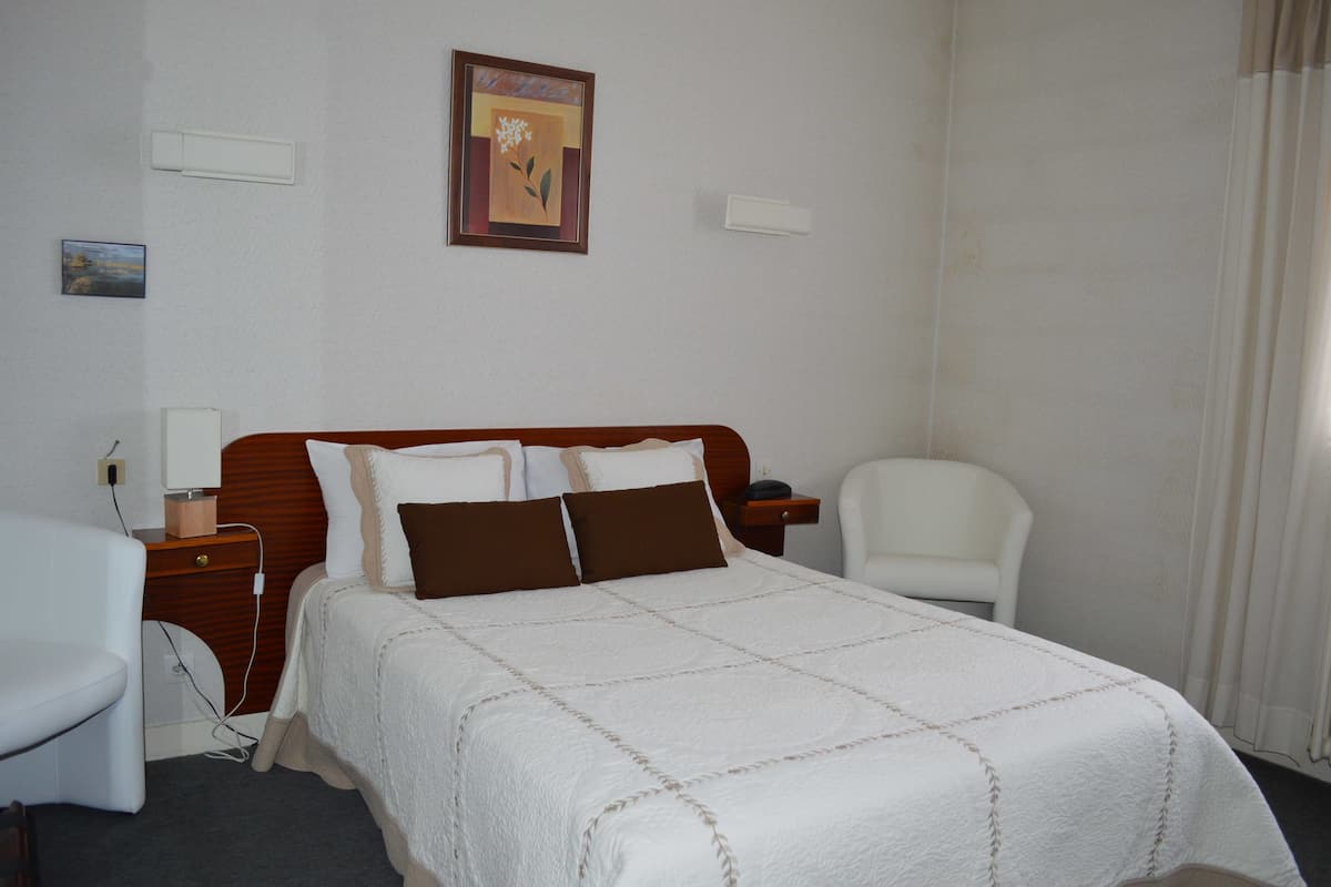 A hotel room with a bed, a bedside table, a chair, a desk, and a painting on the wall.