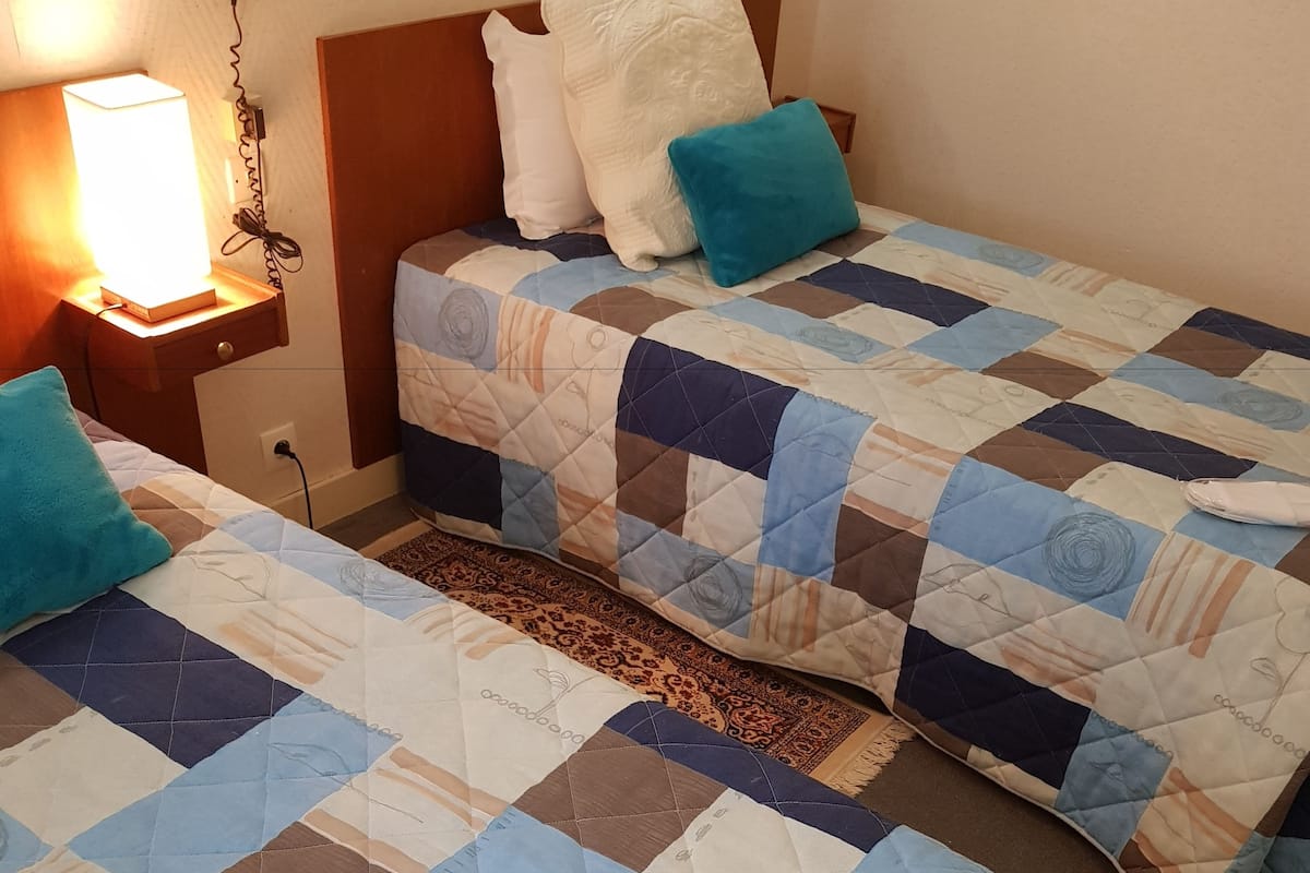 A hotel room with two beds, each with a quilted bedspread featuring geometric patterns and a white pillow. There is a bedside table with a lamp and a phone.