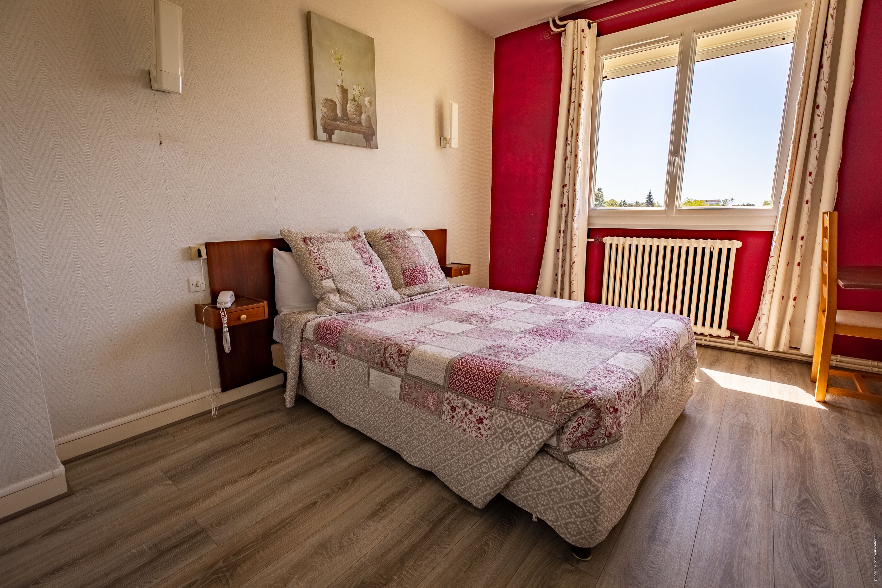 A bedroom with a wooden floor, a bed with a patterned quilt, a nightstand with a phone, a radiator, and a window with curtains.