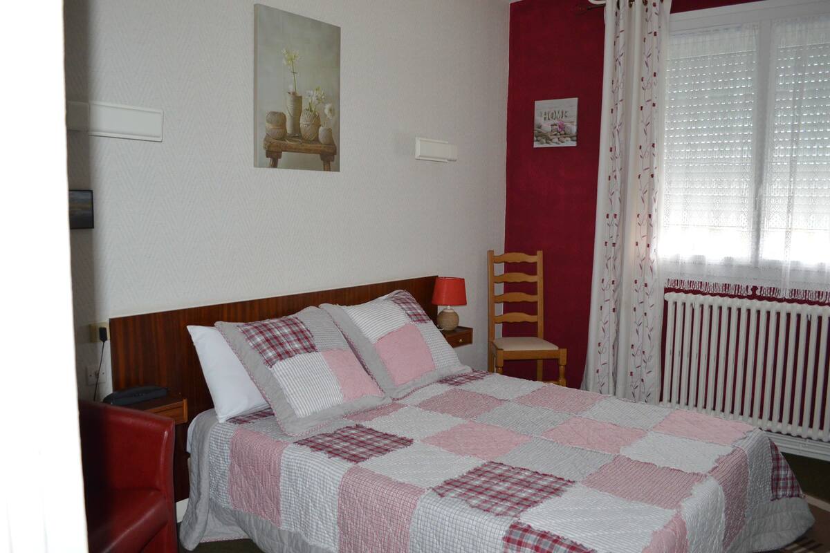 A neatly made bed with a checkered bedspread, a wooden nightstand with a lamp, a red chair, and a window with sheer curtains.