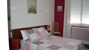Una cama bien tendida con colcha a cuadros, una mesita de noche de madera con lámpara, una silla roja y una ventana con cortinas traslúcidas.