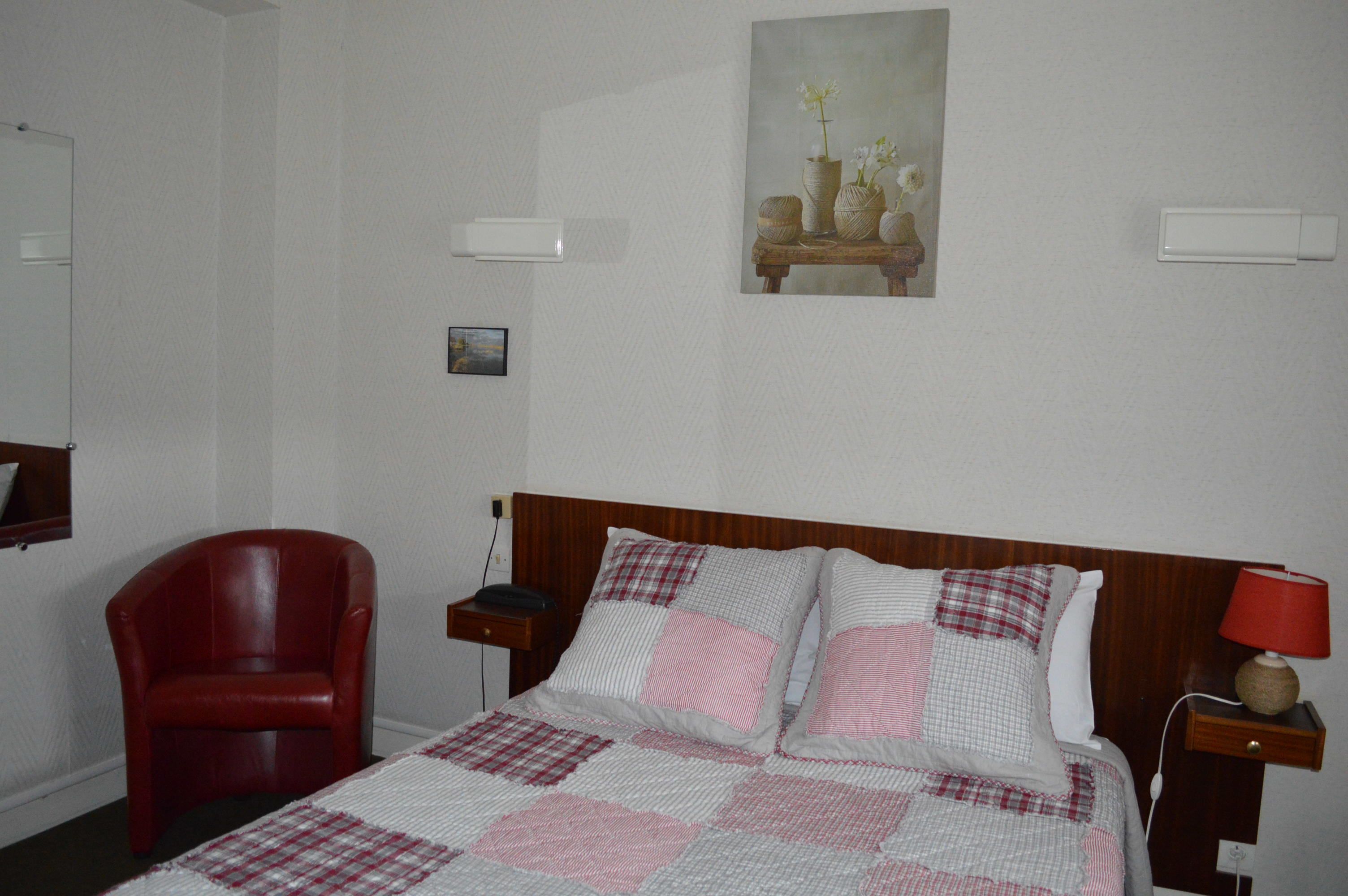 Ein Hotelzimmer mit Bett, einem roten Sessel, einem Nachttisch, einer Lampe und einem Gemälde an der Wand.