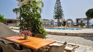 Zona junto a la piscina con una mesa de madera y sillas de mimbre, rodeada de muros de piedra y vegetación.