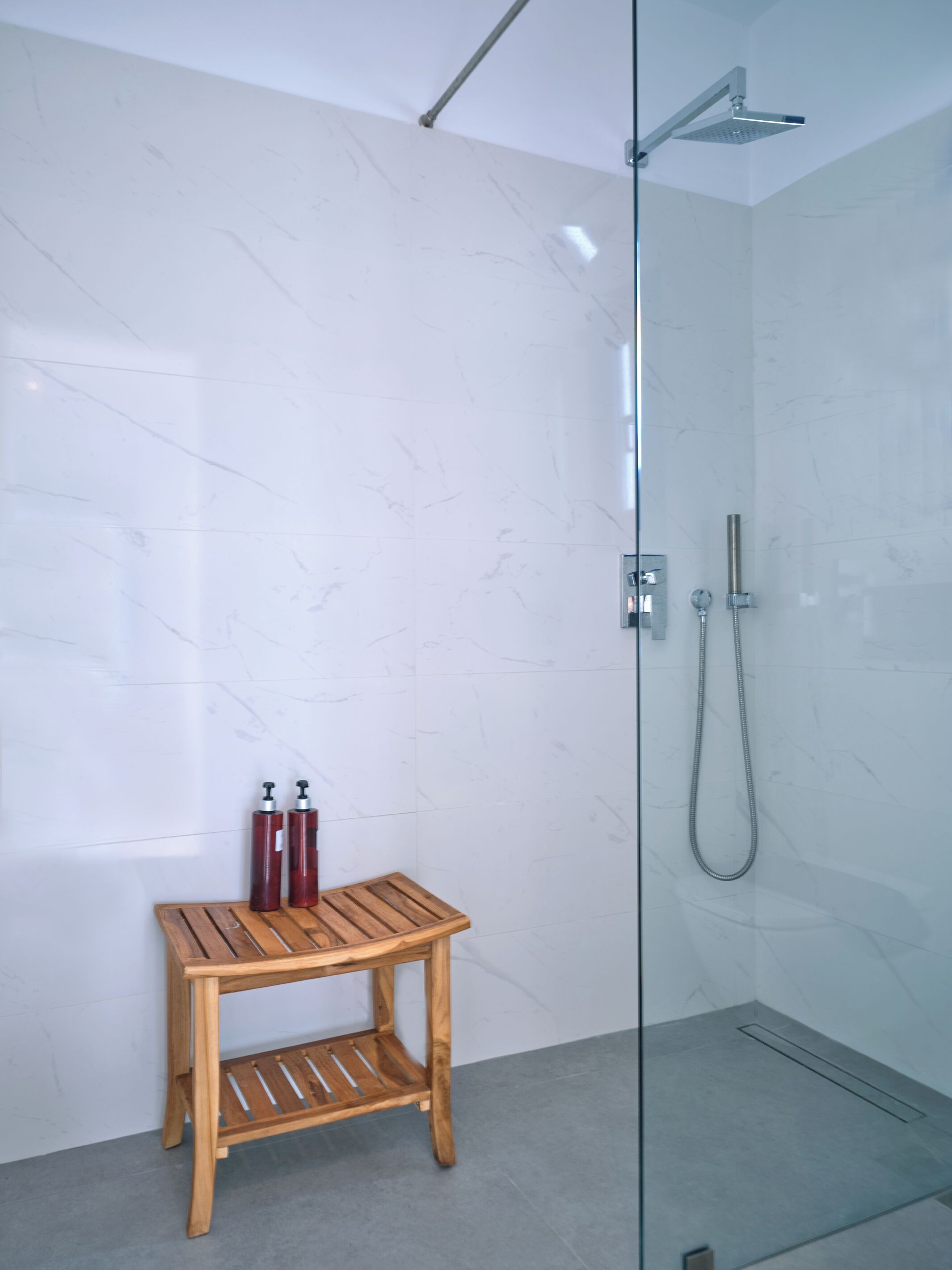 A modern bathroom with a glass shower enclosure, a wooden bench, and two red shampoo bottles.
