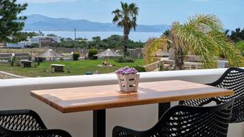 A wooden table with two black chairs, a vase with purple flowers, and a view of a coastal landscape with mountains and palm trees.