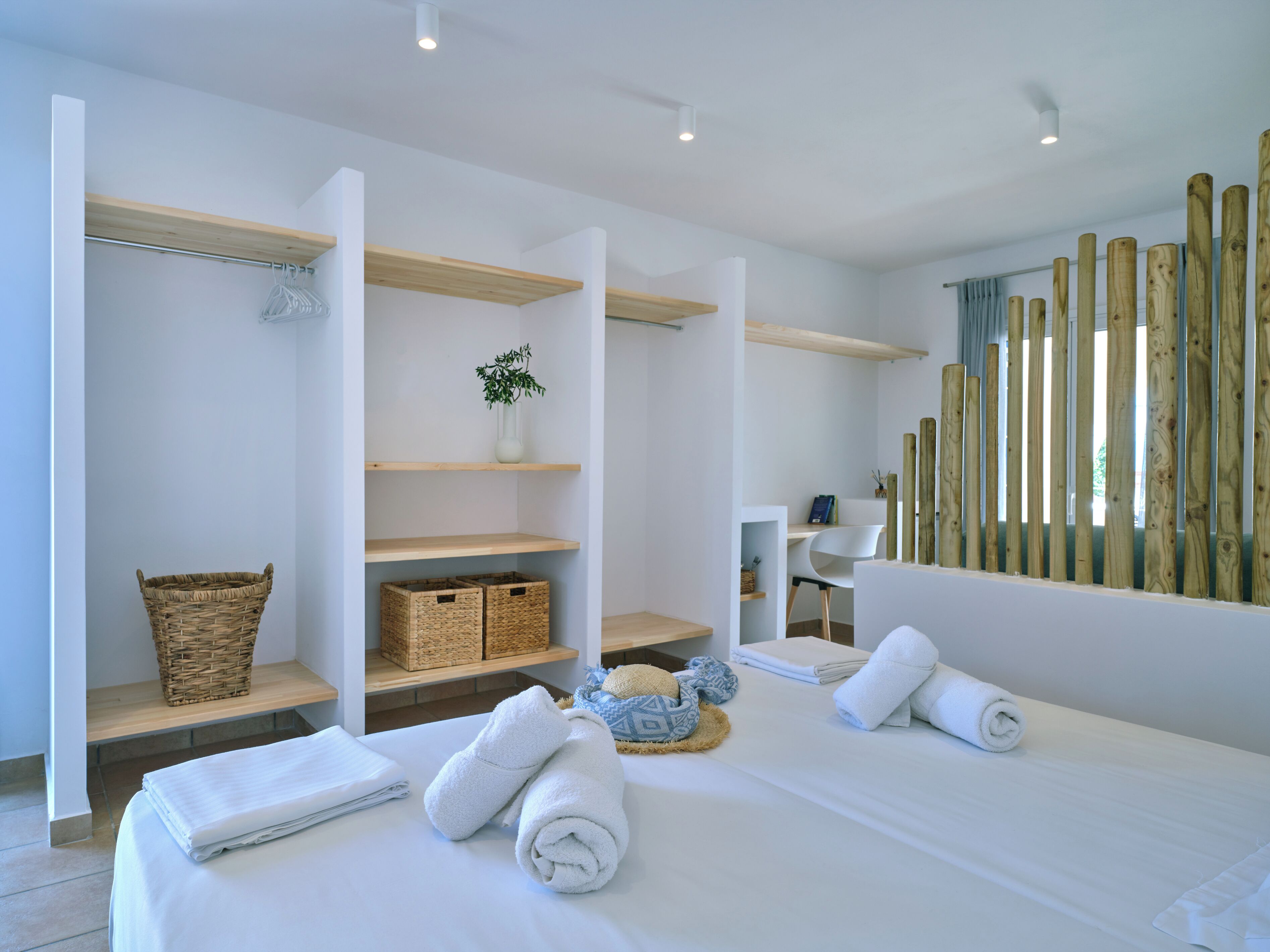 A modern hotel room with a bed, a desk, and a seating area. There are baskets, towels, and a plant on the shelves.