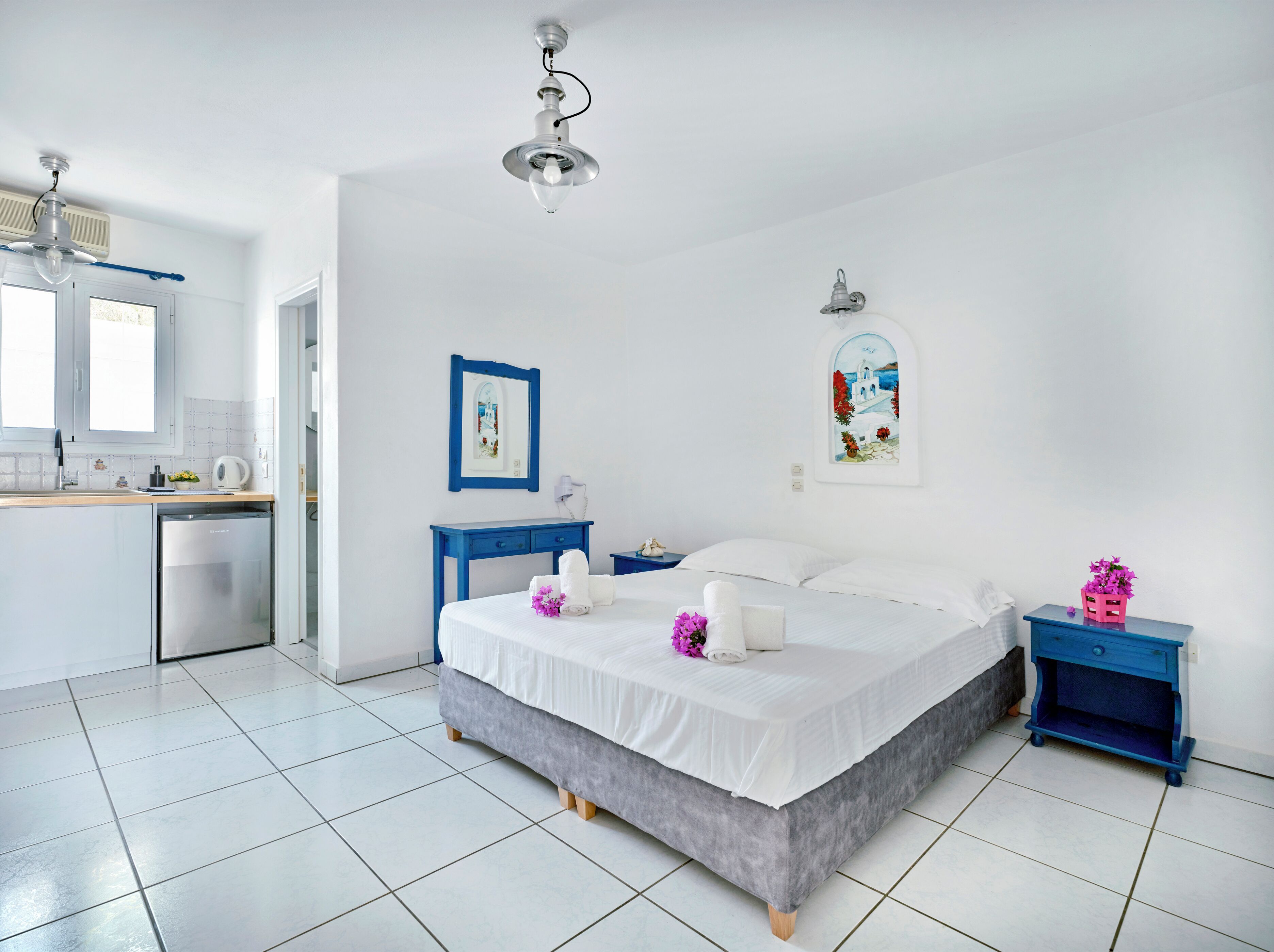 A modern hotel room with a bed, bedside tables, a desk, and a kitchenette.