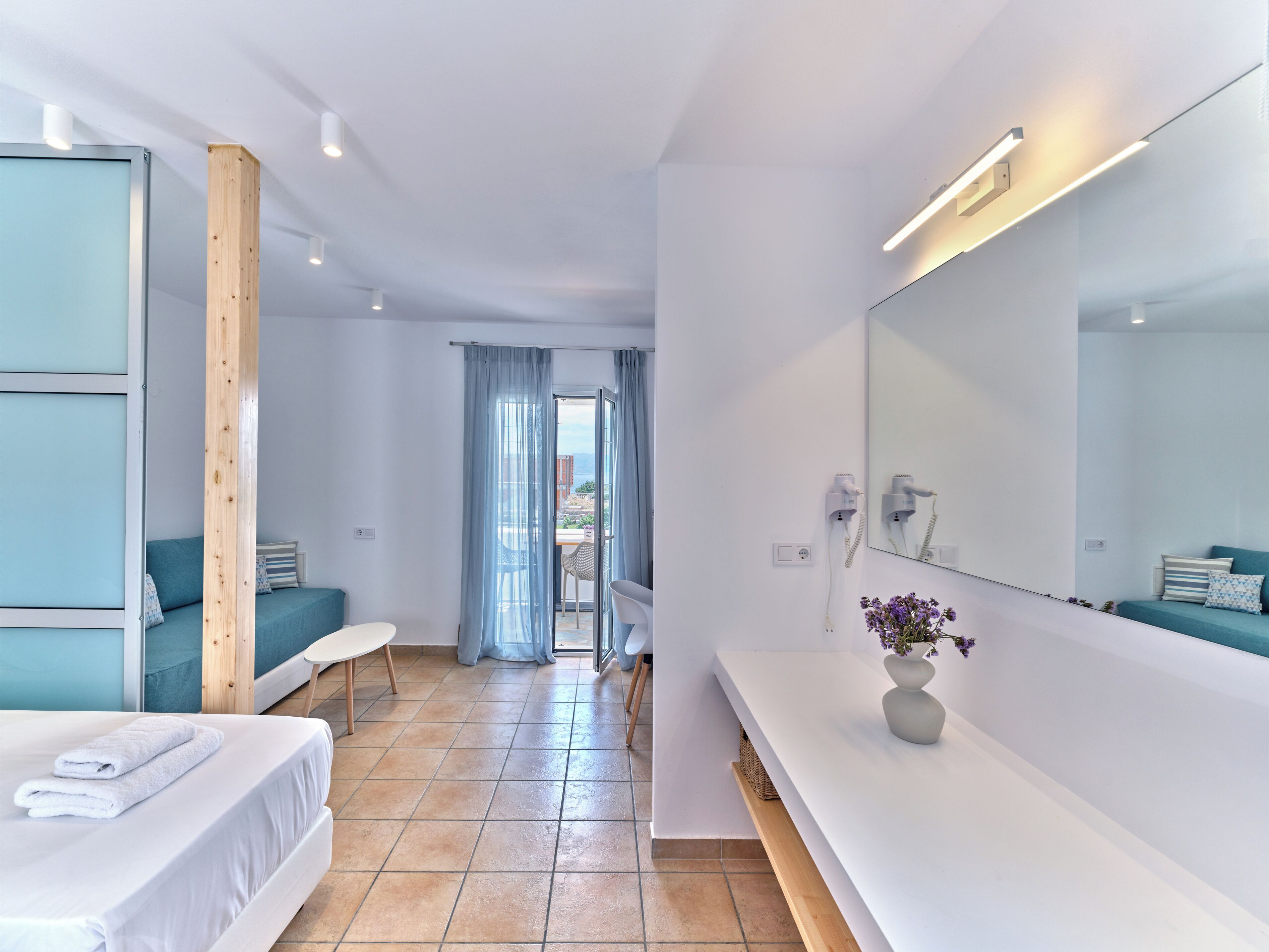 Ein modernes Hotelzimmer mit einem Bett, einer Couch, einem Schminktisch mit Spiegel und Blick ins Freie durch eine Schiebetür.