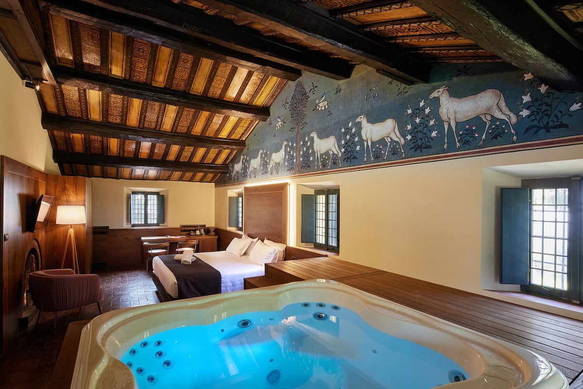 Ein Hotelzimmer mit einer großen Holzbalkendecke, einem Wandbild mit Schafen und Blumen, einer Holzwanne und einem Bett mit weißen Leinen.