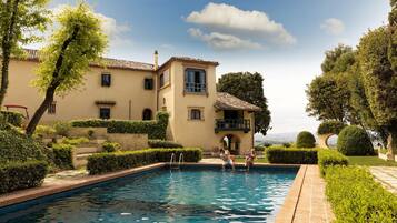 A pool area with a building in the background, surrounded by well-maintained hedges and trees.