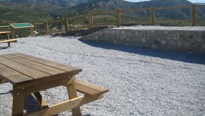 Taula de pícnic de fusta amb bancs sobre un sòl de grava, davant d'una paret de pedra i una barana de fusta, amb vista a un paisatge de muntanya.