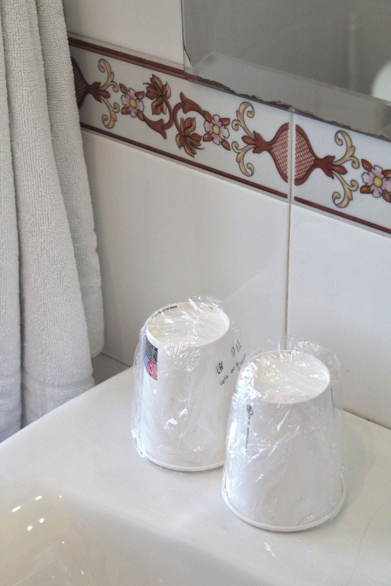 A bathroom with a white sink, two cups wrapped in plastic, and a decorative tile border.