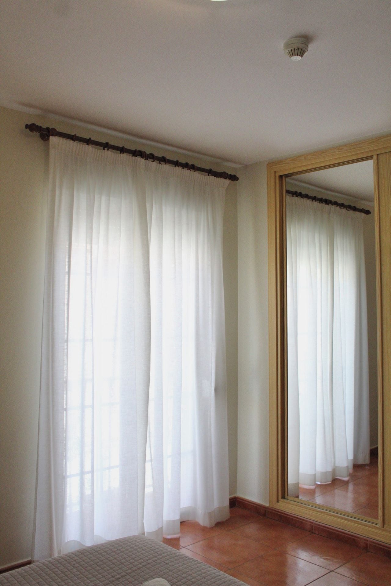 A room with a large mirror reflecting curtains, a window with sheer curtains, and a tiled floor.