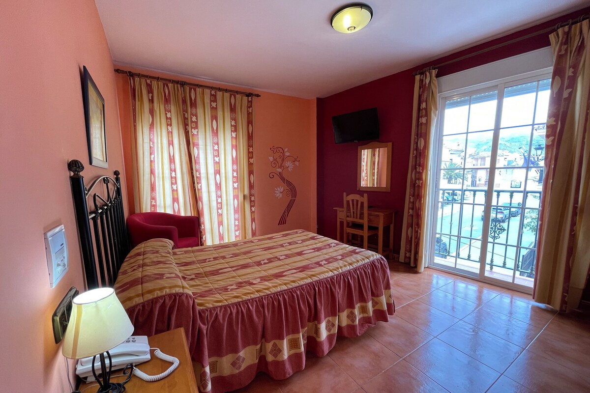 Quarto de hotel com cama, escrivaninha, cadeira, televisão e janela com vista para o exterior.
