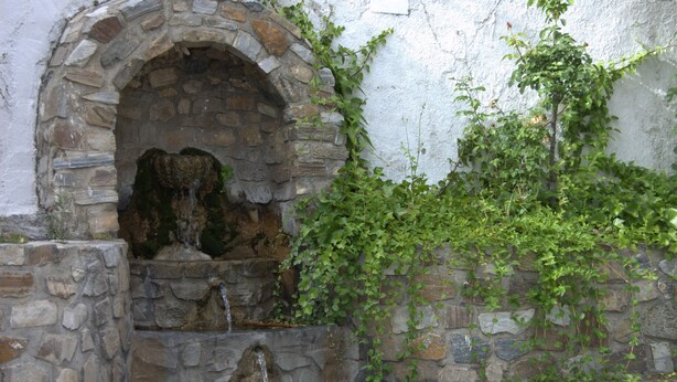 Una font de pedra amb aigua corrent, envoltada per un mur de pedra i vegetació.
