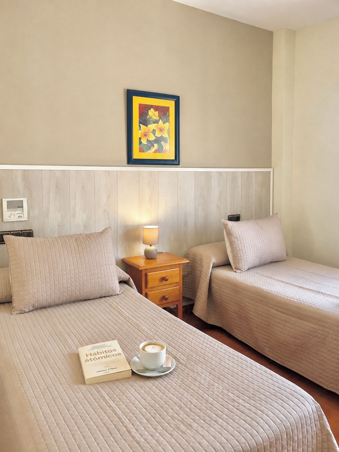 Habitación de hotel con dos camas, una mesita de noche con lámpara, un cuadro enmarcado en la pared y un libro y una taza sobre la cama.