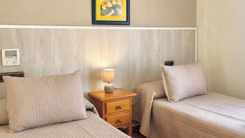 Habitación de hotel con dos camas, una mesita de noche con lámpara, un cuadro enmarcado en la pared y un libro y una taza sobre la cama.