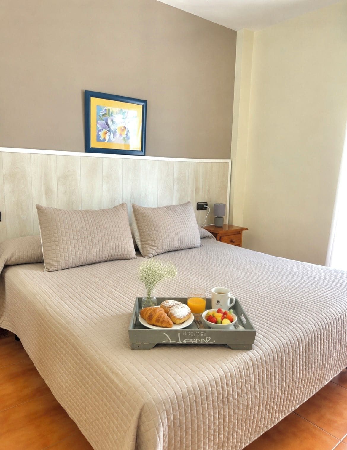 Quarto de hotel com uma cama grande, uma mesa de cabeceira com abajur, um quadro na parede e uma bandeja com itens de café da manhã.