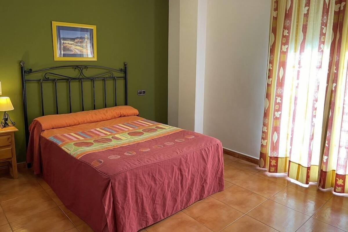 Une chambre à coucher avec un lit, une table de chevet, une lampe, un tableau encadré au mur et un rideau.