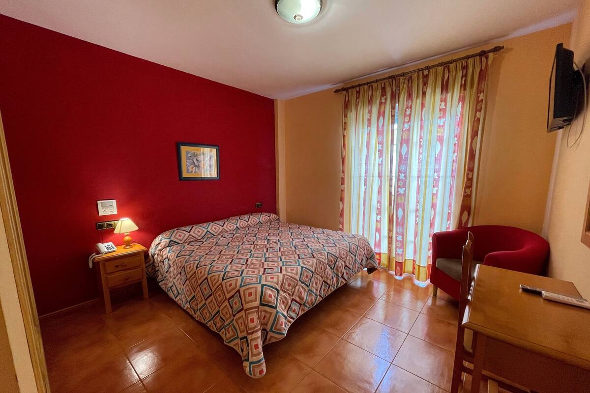 Une chambre à coucher avec une couvre-lit à motifs, une lampe de chevet, un téléphone, une chaise et un téléviseur fixé au mur.