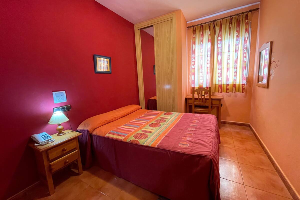 Une chambre à coucher avec un lit, une table de chevet avec une lampe, une chaise, un bureau et une fenêtre avec des rideaux.