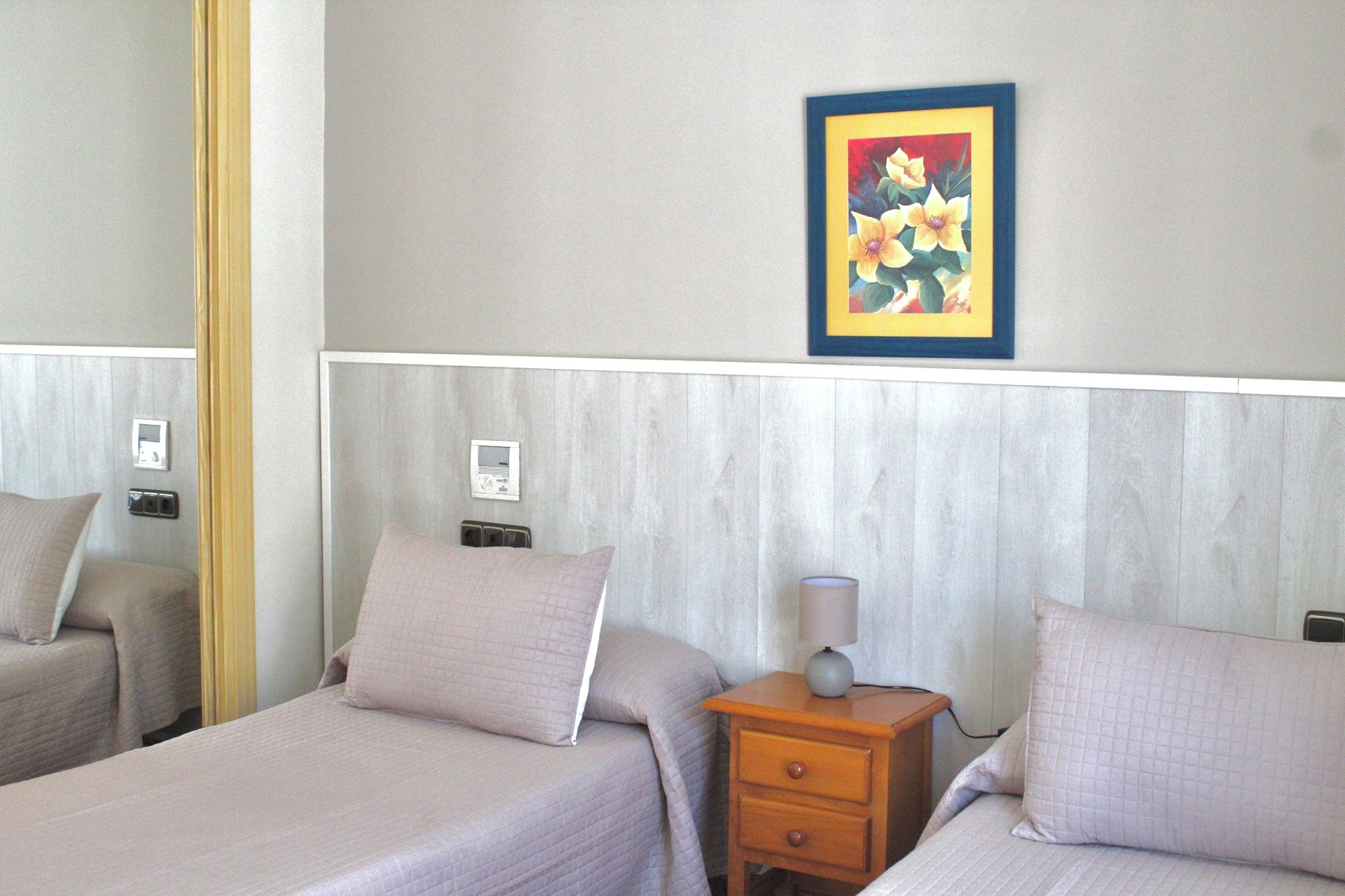 A hotel room with two beds, a bedside table with a lamp, a wall-mounted thermostat, and a framed picture on the wall.
