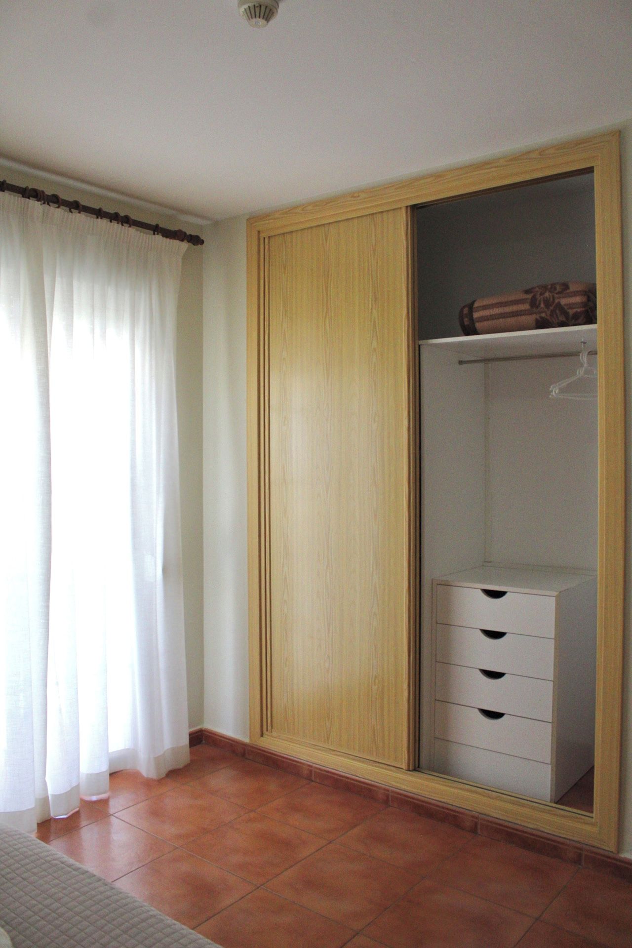 Una habitación con un armario de madera, un aparador blanco y una ventana con cortinas blancas.