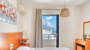 Quarto de hotel com uma cama, mesa de cabeceira com luminária, cômoda, espelho e vista para o mar.