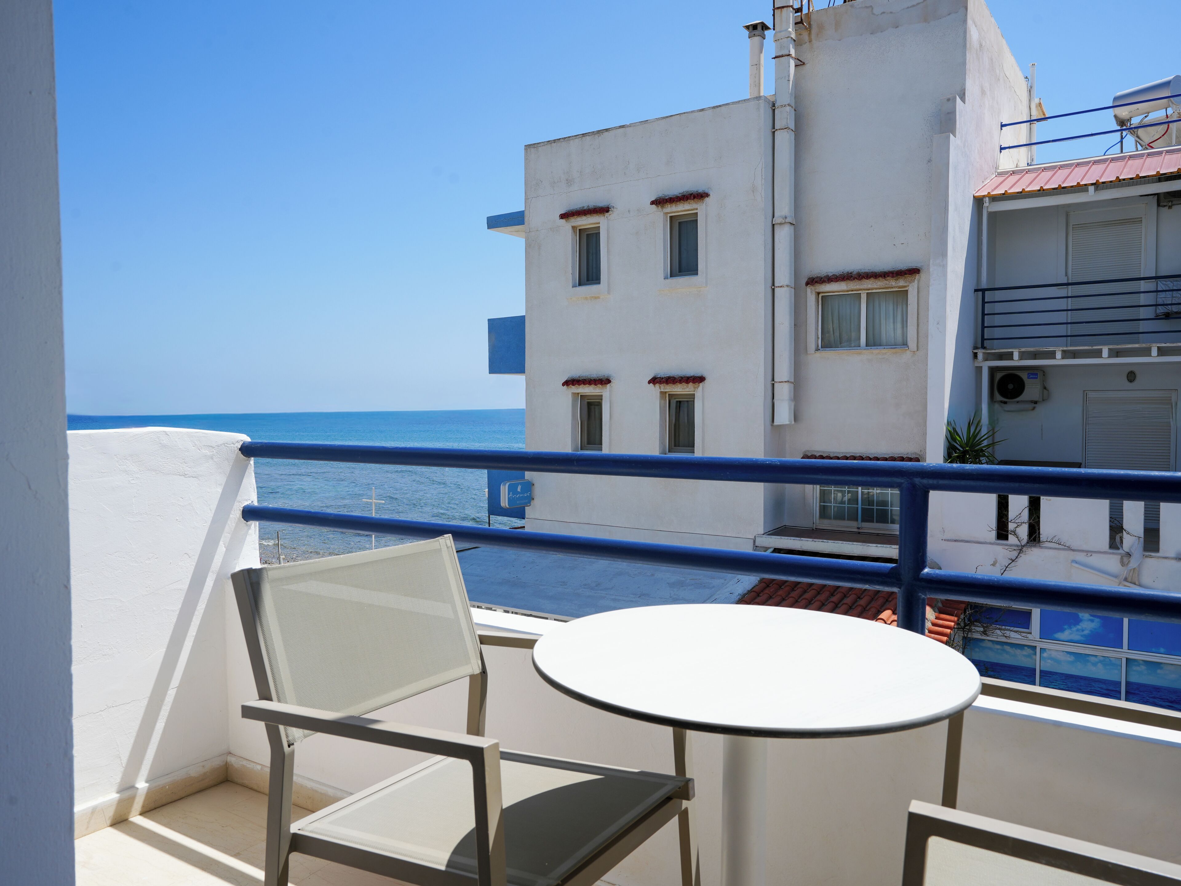 Un balcón con una mesa y sillas blancas, con vista al mar azul y edificios de techos rojos.