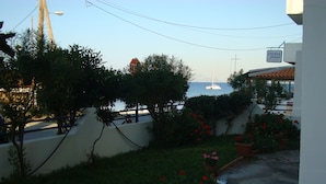 Vista costeira com um barco ao longe, um letreiro para 'ANEMOS Restaurant' e um jardim com plantas em vasos.