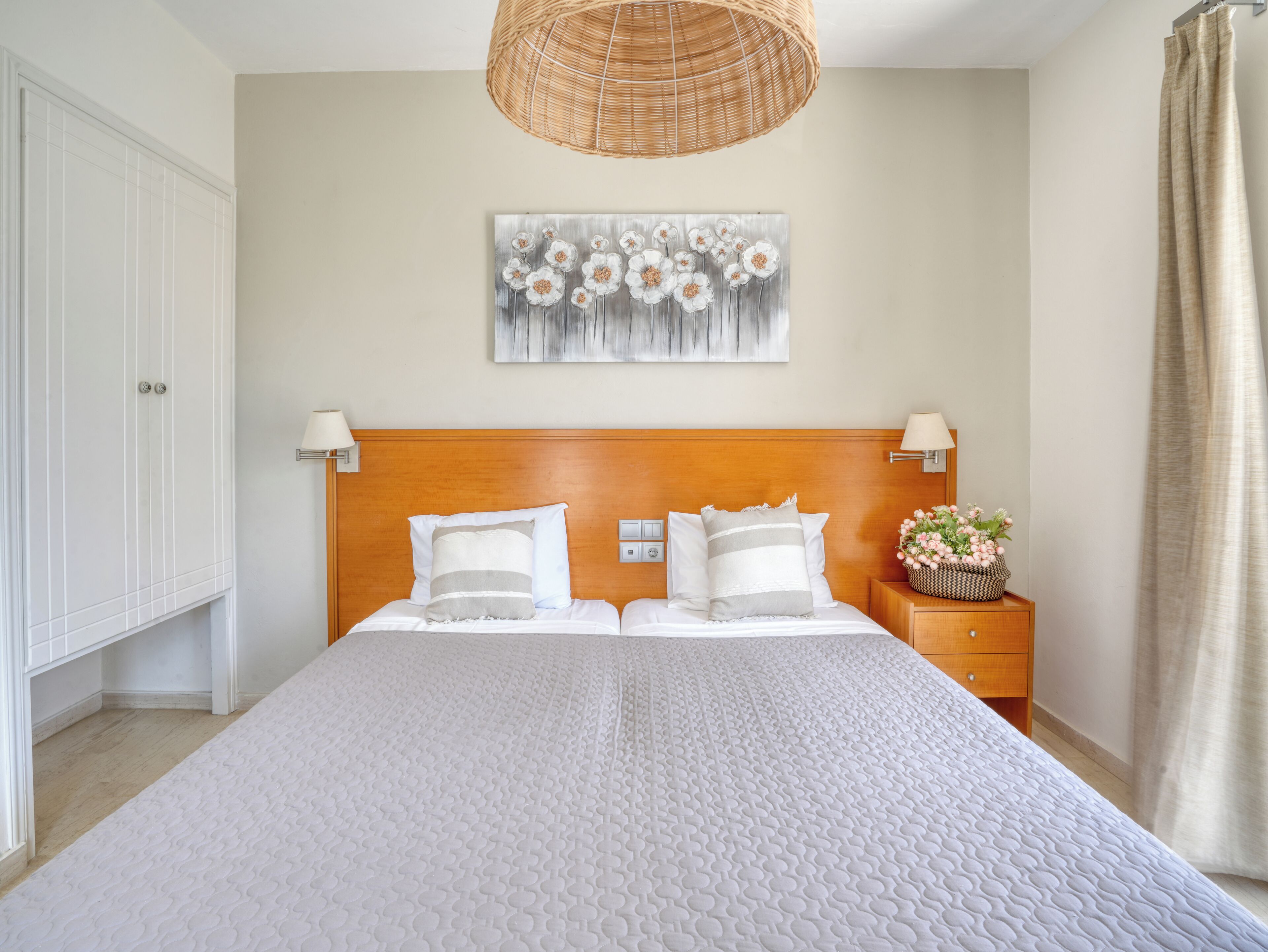 A bedroom with a large bed, a wooden headboard, a wall-mounted artwork, bedside lamps, and a nightstand with a basket of flowers.