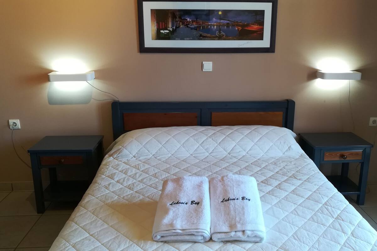 A neatly made bed with a quilted white duvet, two bedside tables with lamps, a framed picture on the wall, and a small nightstand with a drawer.