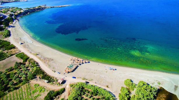 An aerial view of a coastal area with a beach, clear waters, and surrounding vegetation.