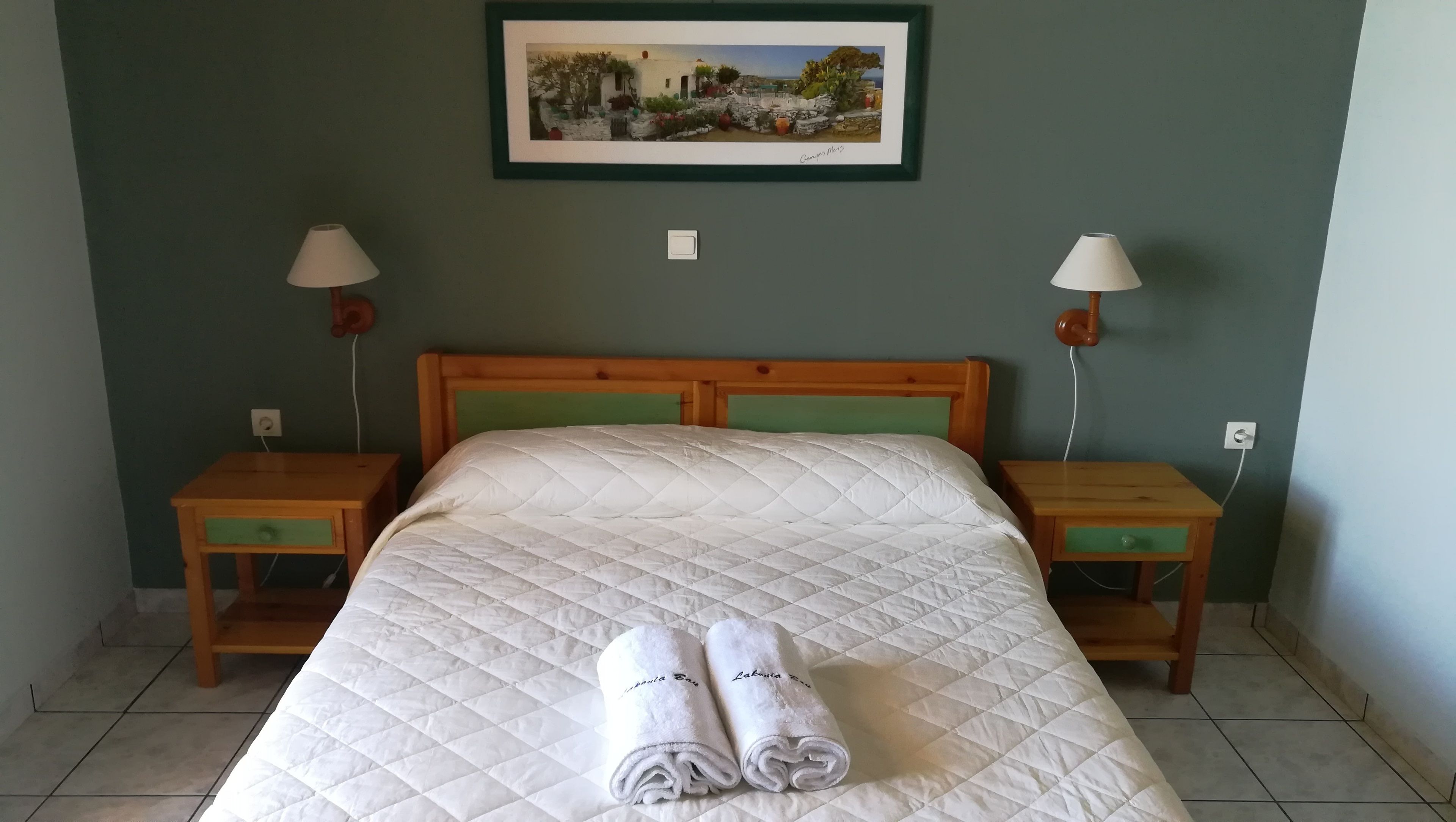 Un lit bien fait, recouvert d’une couette blanche matelassée, deux tables de chevet en bois avec des lampes, un tableau encadré au mur et un sol carrelé.
