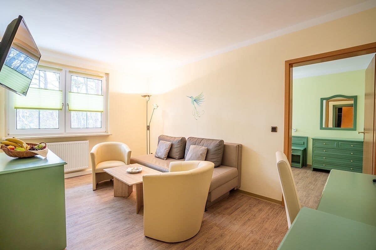 Uma sala de estar moderna com um sofá, duas poltronas, uma mesa de centro e uma TV fixada na parede. Há uma janela com persianas, um abajur e um objeto decorativo na parede.