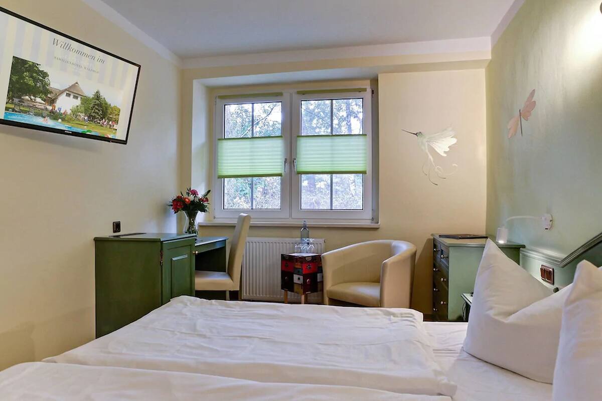 Quarto de hotel com cama, escrivaninha, cadeira, radiador e janela com persianas.