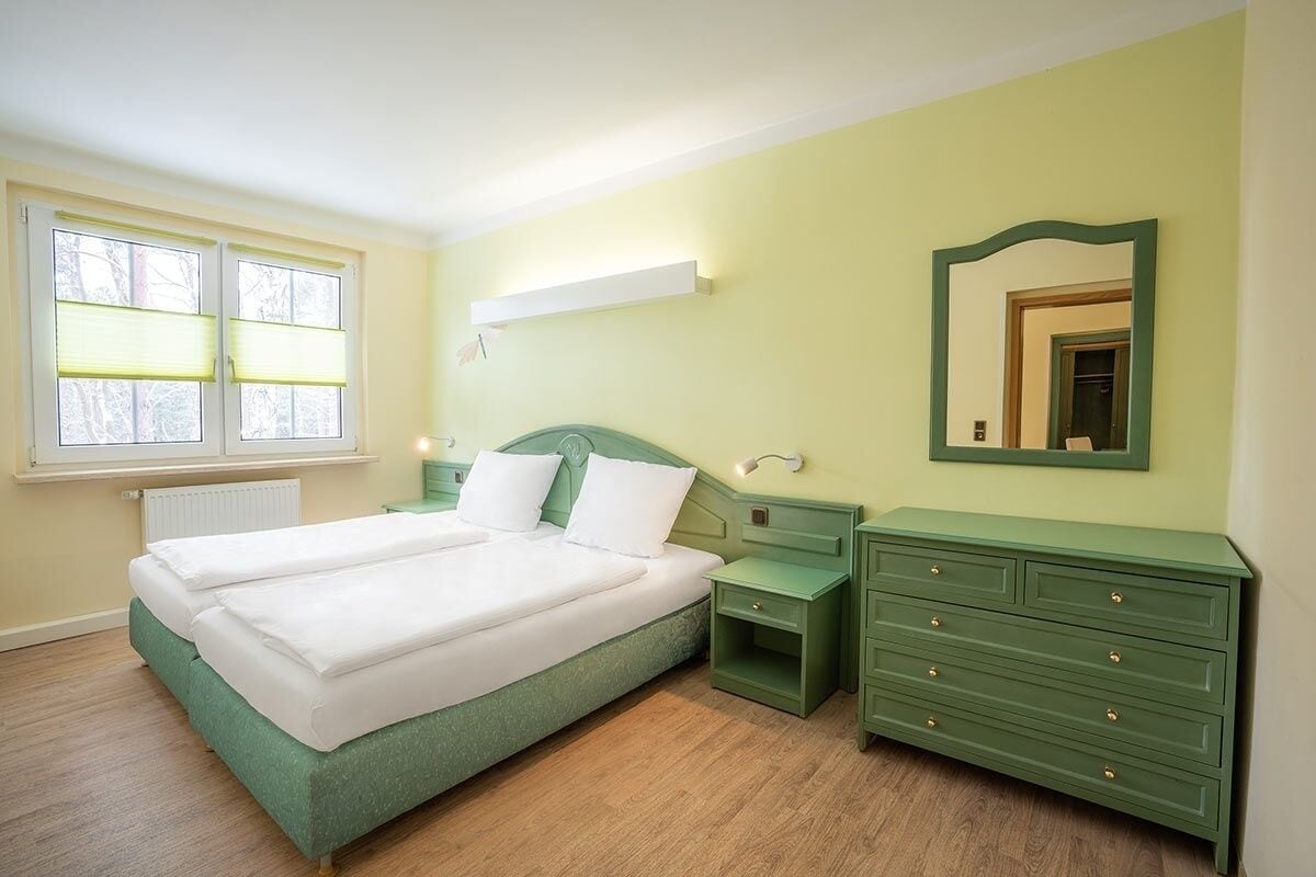Quarto com uma cama de casal, mesas de cabeceira, cômoda, espelho e janela com persianas.
