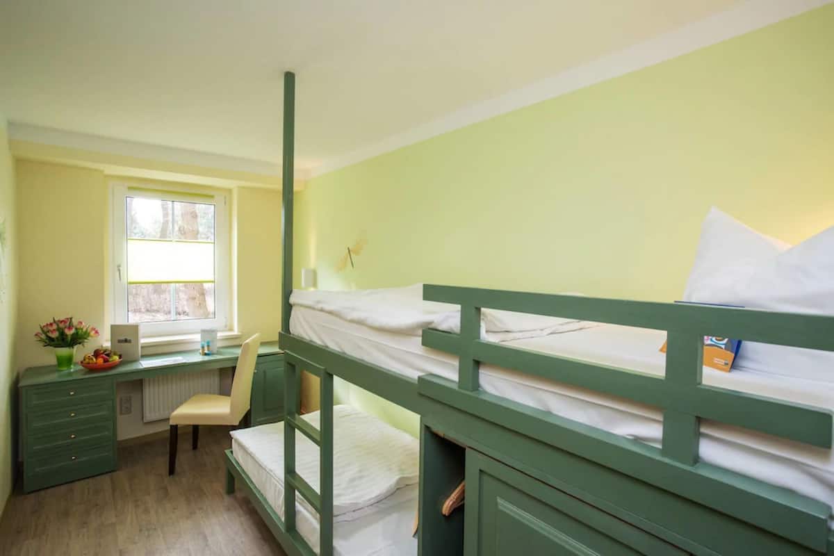 Quarto com beliche, escrivaninha, cadeira, radiador e janela com cortinas.